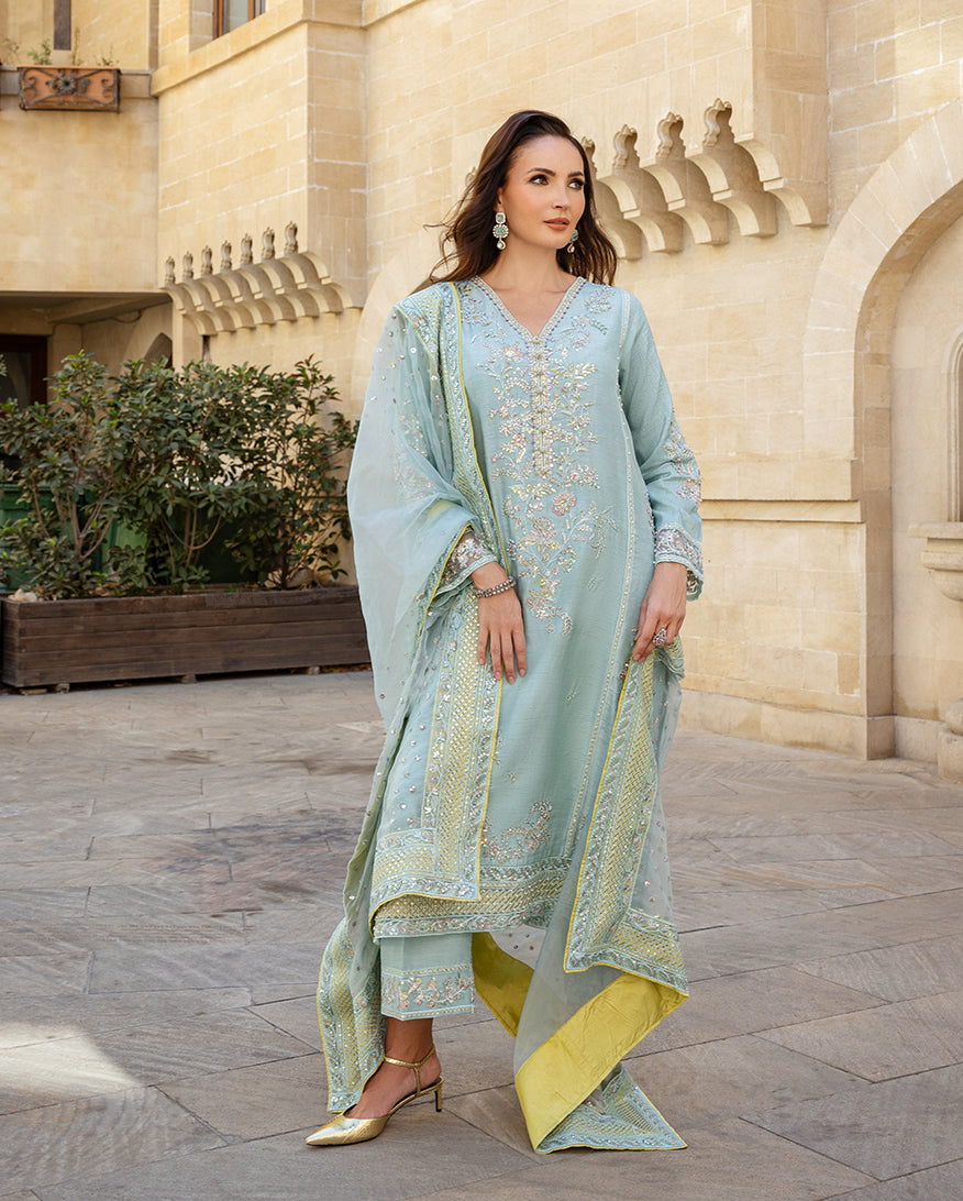 Woman wearing icy blue raw silk suit with organza dupatta blowing in sandstone courtyard
