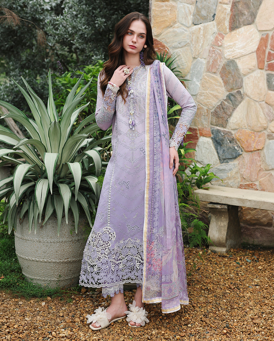 Woman wearing lavender embroidered lawn unstitched suit with lace hem, standing near stone wall, Secret Garden collection